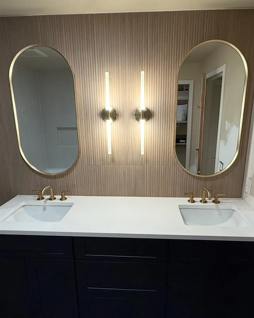 Double vanity with quartz countertop, brushed gold faucets, oval mirrors and LED sconces on fluted wall tile